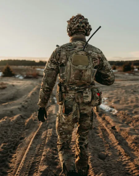 Giocatore di spalle in mimetica multicam, con zaino tattico e antenna radio, su un terreno aperto al tramonto